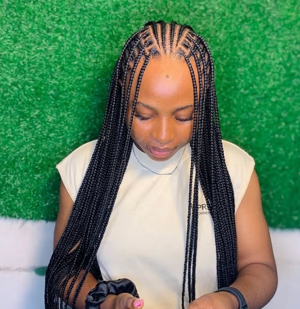 A woman wearing flip-over Fulani braids, showcasing a combination of cornrows and long knotless braids. The background is a green grass-like texture.