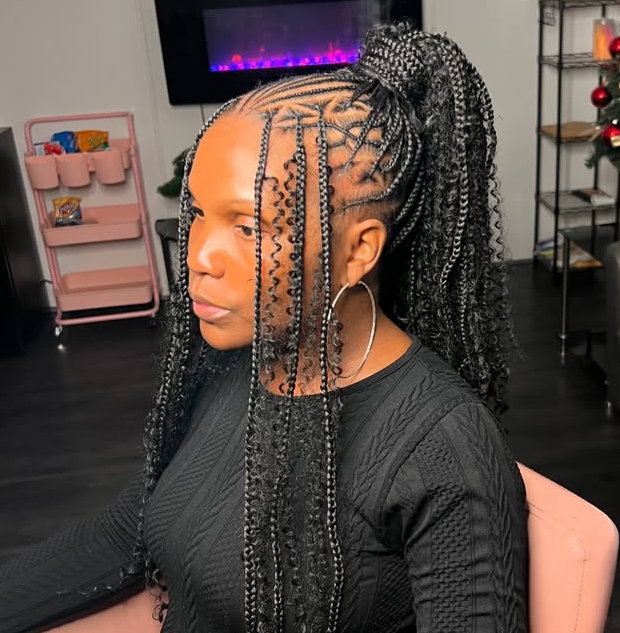 A woman with flip-over Fulani braids styled in a high ponytail, featuring intricate cornrow patterns on the sides and wavy ends. She is seated in a cozy setting with a modern decor.