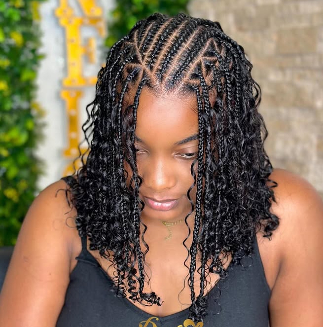 Close-up of a woman with a flip-over Fulani braids hairstyle featuring intricate cornrows at the top and curly ends. The background includes green foliage, highlighting the stylish look.