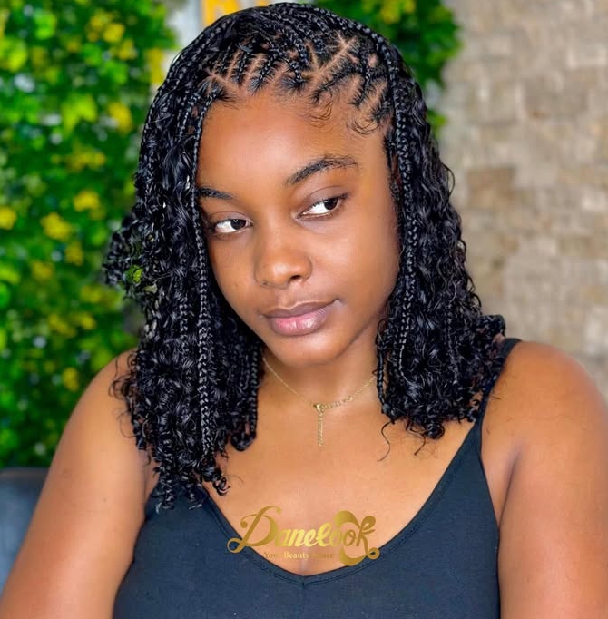 A close-up of a person with flip-over Fulani braids hairstyle, featuring a blend of cornrows and loose curls. The background is softly blurred with greenery.