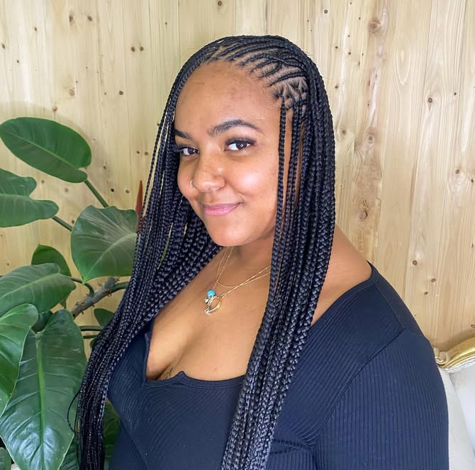 A woman with long flip-over Fulani braids hairstyle, showcasing intricate cornrows on the sides and knotless braids flowing down, standing in front of a wooden wall with plants in the background.