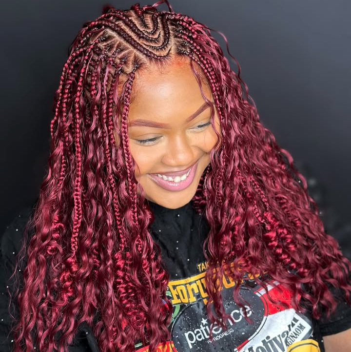 A woman with red flip-over Fulani braids, showcasing a mix of knotless braids and cornrows, smiling while looking at the side.
