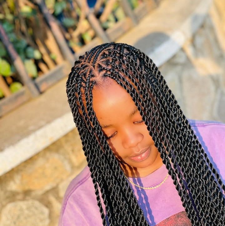A person with long black flip-over Fulani braids is looking down, showcasing the intricate braid pattern on their head. They are wearing a purple shirt and a thin necklace against a softly blurred outdoor background.