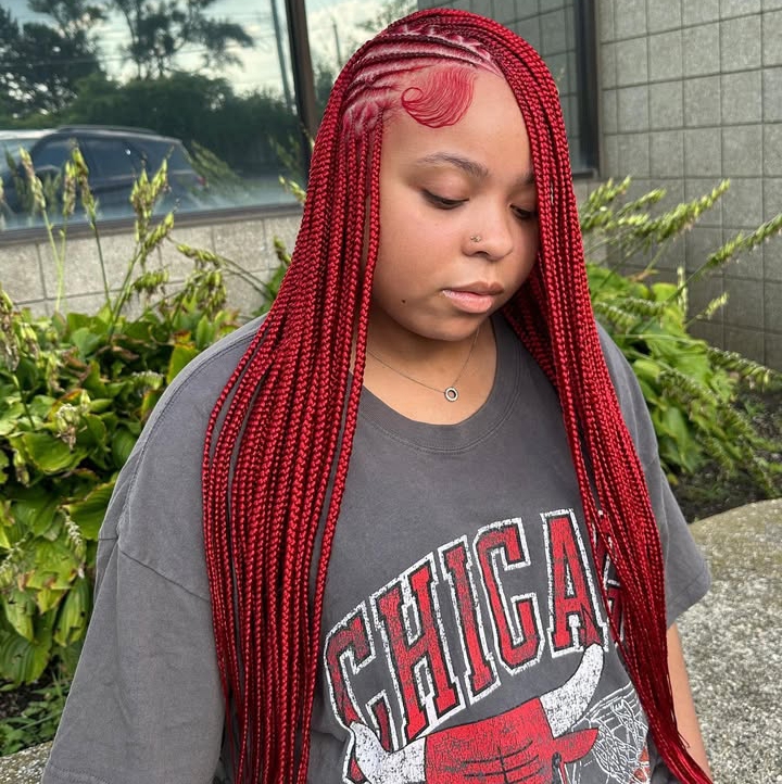 A close-up of a person with long red flip-over Fulani braids, featuring intricate cornrow patterns at the front, wearing a loose graphic t-shirt, against a backdrop of greenery.