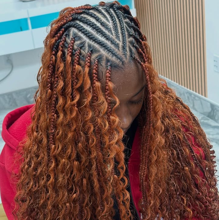 A woman with flip-over Fulani braids featuring a blend of knotless braids and cornrows. The hairstyle has an orange color with curly ends and is styled with the front cornrows in a triangular pattern. The setting appears to be a salon.