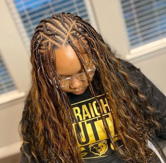 A young woman wearing flip-over Fulani braids with wavy ends, sitting in a chair, wearing glasses and a black sweatshirt with a golden design.