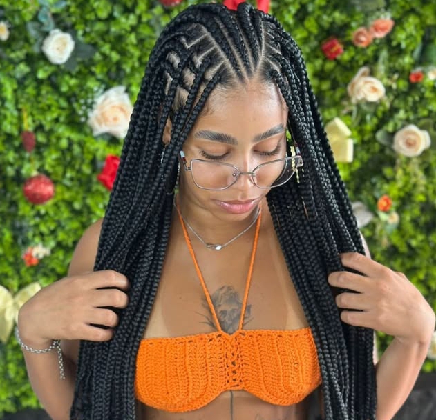 A woman wearing flip-over Fulani braids, styled with long, straight hair and glasses, standing in front of a green floral backdrop, while wearing an orange top.