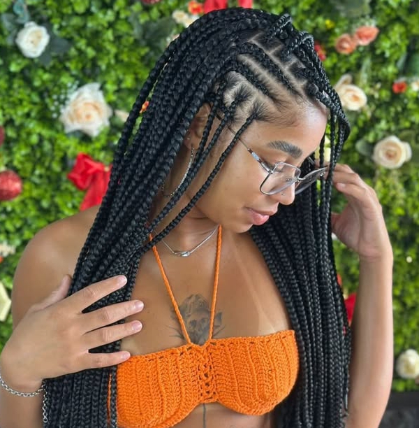 A woman showcasing a stylish flip-over Fulani braids hairstyle, featuring knotless box braids and cornrows, against a lush floral background. She is wearing an orange crochet top and glasses.