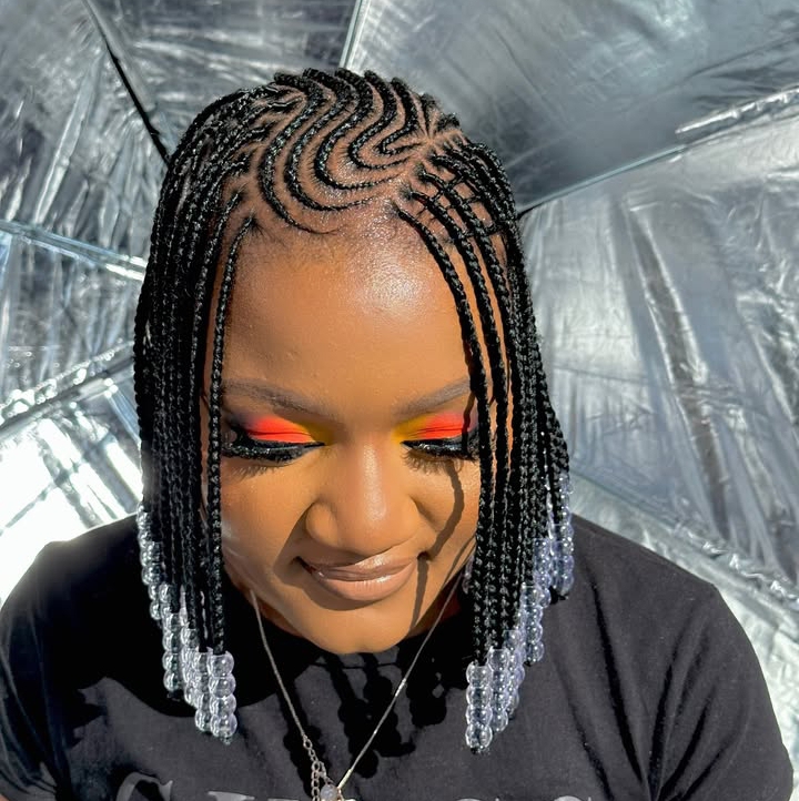 Close-up of a woman with flip-over Fulani braids styled into intricate patterns, featuring colorful makeup and bead decorations on the ends.