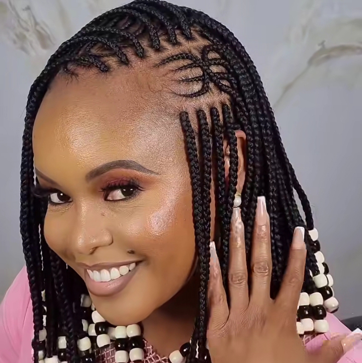 A woman displaying a beautiful flip-over Fulani braids hairstyle, featuring knotless braids and cornrows, adorned with beads, smiling and showing off her manicure.