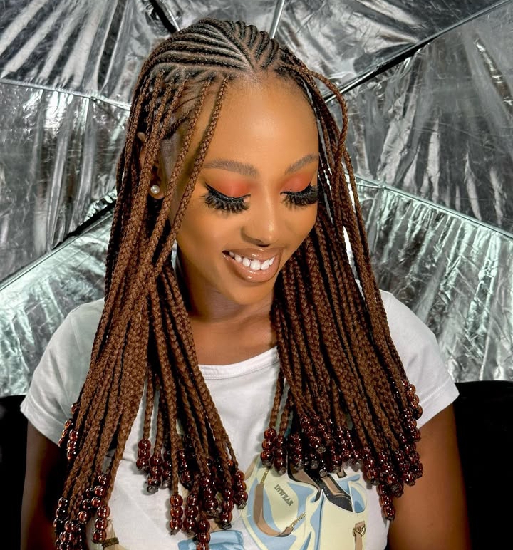 A woman with flip-over Fulani braids decorated with beads, smiling while wearing a graphic t-shirt.