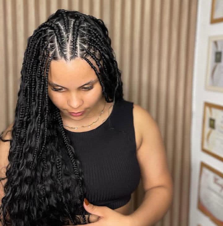 A woman with long, black flip-over Fulani braids is looking down, wearing a black crop top. The hairstyle features knotless braids and cornrows, showcasing a braided pattern along the sides and back.
