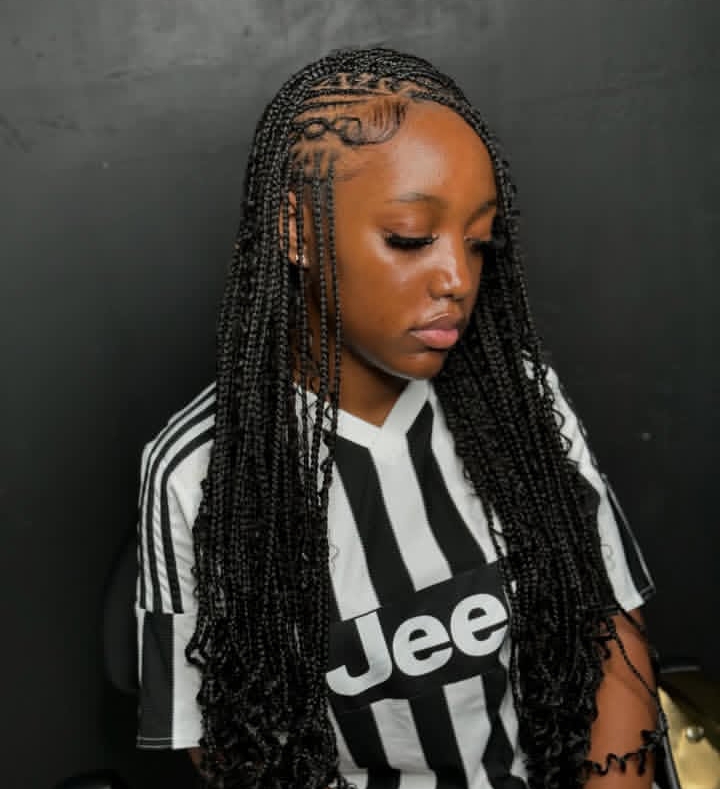 A young woman wearing a black and white striped jersey styled with long flip-over Fulani braids, showcasing intricate cornrow patterns along her hairline.