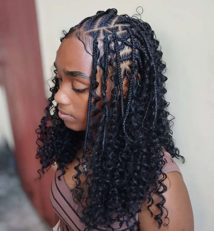 A woman with flipped-over Fulani braids hairstyle featuring knotless braids at the back and cornrows on the sides. Her hair is styled with curly ends, and she is wearing a sleeveless top, facing slightly to the side.