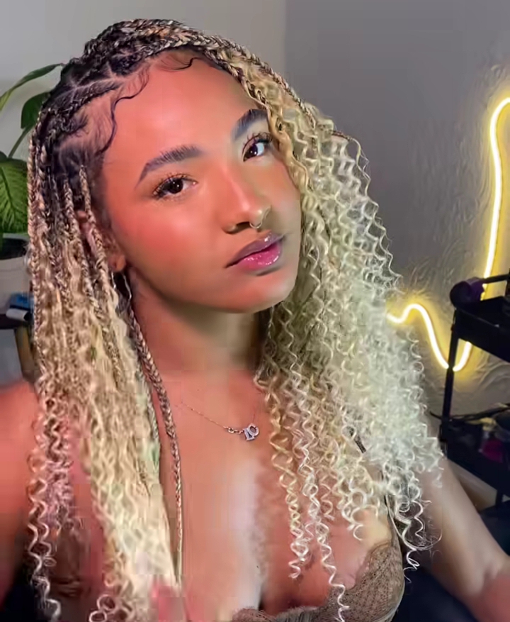 Young woman with flip-over Fulani braids hairstyle, featuring blonde curls and a stylish look, sitting indoors with a softly lit background.