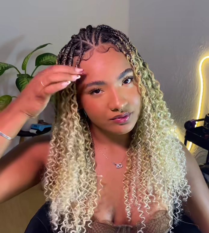 A woman with blonde flip-over Fulani braids hairstyle, styled with curly ends, touches her hair while posing in a well-lit room.