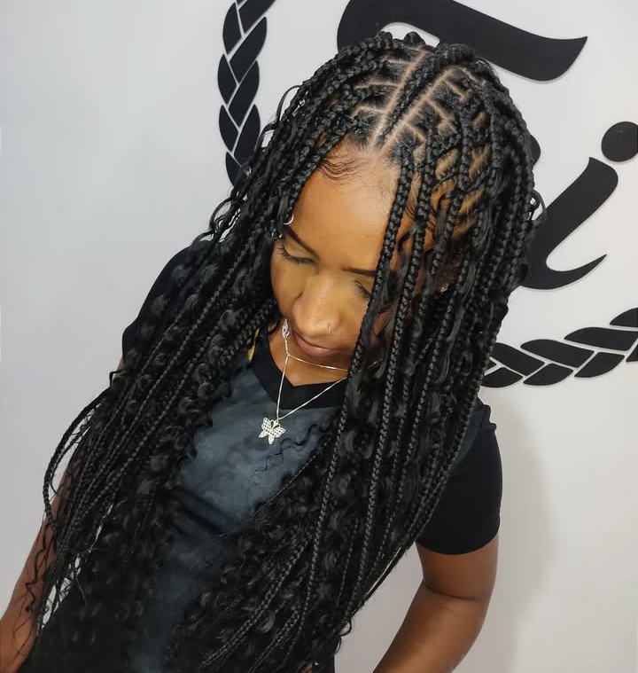 A woman with long flip-over Fulani braids hairstyle, featuring knotless braids and cornrows, standing in front of a neutral background with a stylish logo.